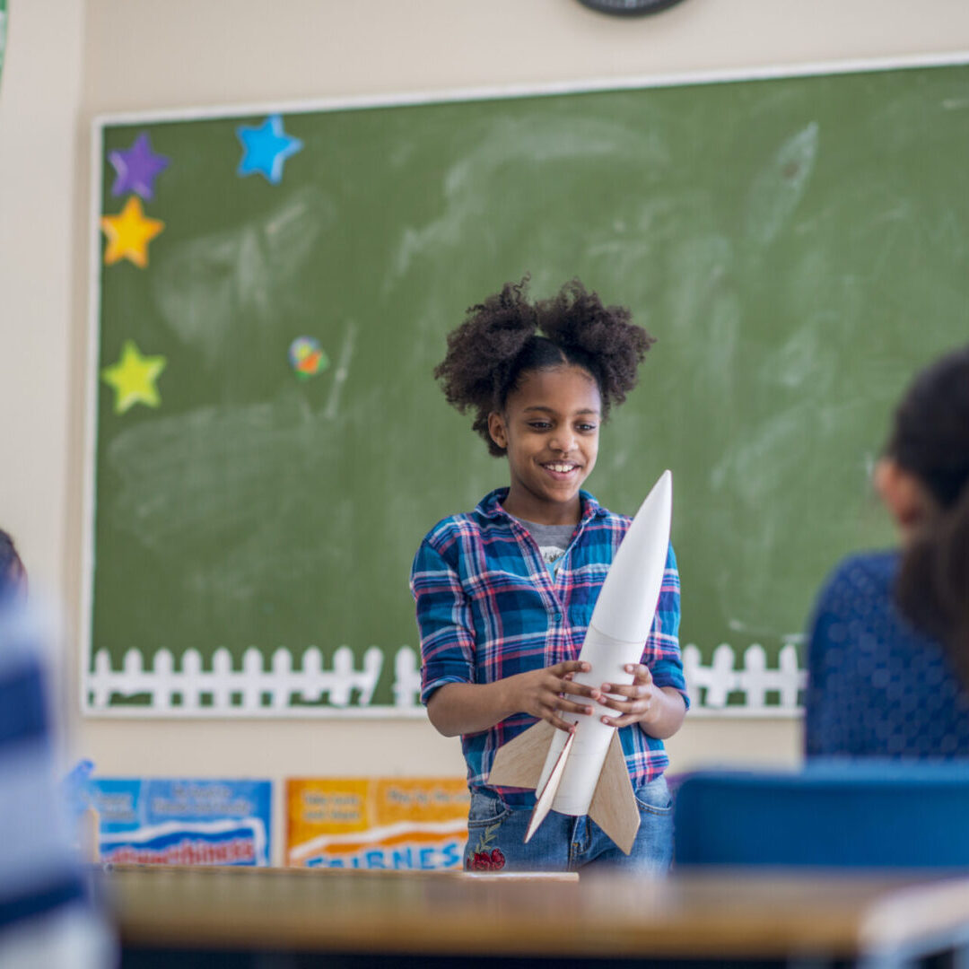 Young girl proudly holds a model rocket in a classroom.